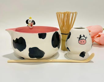 May include: A ceramic matcha tea set featuring a cow print design. The set includes a bowl with a dog figurine, a whisk, a cup, and a bamboo scoop. The bowl has a pink interior and black spots. The cup has a cow face.