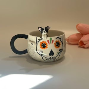 May include: A ceramic mug with a skull design, featuring orange flower accents and a black handle. A small black and white dog figurine is perched on the rim. The mug has a speckled, off-white finish.