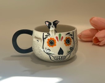 Puede incluir: Una taza de cerámica con diseño de calavera, con detalles de flores naranjas y un asa azul oscuro. Una pequeña figura de perro blanco y negro está posada en el borde. La taza tiene un acabado moteado blanquecino.