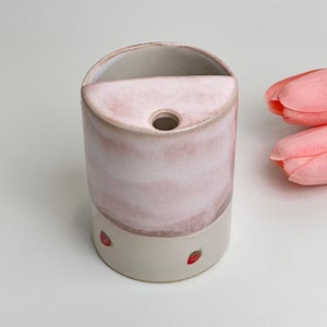 Peut inclure: Un vase en céramique avec un effet aquarelle rose clair et une base beige. Le vase a un dessus semi-circulaire et un petit trou. De petites fraises rouges sont peintes sur la base. Des tulipes roses sont en arrière-plan.