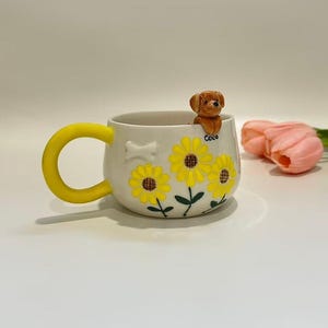 May include: A cream-coloured ceramic mug with a yellow handle, adorned with yellow sunflowers and green leaves. A small brown dog figurine sits on the rim. The word "Coco" is written on the mug.