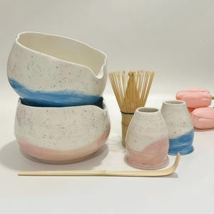 May include: A ceramic matcha set featuring two speckled bowls with blue and pink painted bases, two matching small cups, a bamboo whisk, and a bamboo scoop. The bowls have a pouring spout, and the set is accompanied by two soft pink artificial tulips.