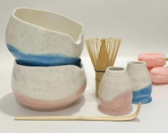 May include: A ceramic matcha set featuring two speckled bowls with blue and pink painted bases, two matching small cups, a bamboo whisk, and a bamboo scoop. The bowls have a pouring spout, and the set is accompanied by two soft pink artificial tulips.