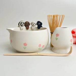 May include: A ceramic matcha bowl with pink tulip patterns and four small dog figurines perched on the rim, accompanied by a bamboo whisk, a bamboo scoop, and a white ceramic whisk holder decorated with a matching pink tulip design.