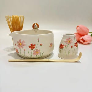 Könnte beinhalten: Ein cremefarbenes Matcha-Schüssel-Set aus Keramik mit einer Hundefigur, Blumendesign und Bambusbesen. Das Set enthält eine Schüssel, eine kleine Vase, einen Bambuslöffel und einen Schneebesen. Rosa Blumen und grüne Stiele verzieren die Schüssel und die Vase.