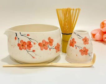 Op de afbeelding: Een keramische matchakom en een bijpassende gardehouder, beide met een crèmekleurige basis, handgeschilderde oranje kersenbloesems en zwarte takken. Een bamboe garde en een lange, slanke bamboe theeschep zijn in de set inbegrepen.