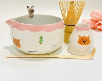May include: A ceramic matcha bowl with a pink scalloped rim, a small cat figurine on the edge, and bear, tree, and fish illustrations. Includes a bamboo whisk, a bamboo scoop, and a ceramic whisk holder with a bear face illustration.