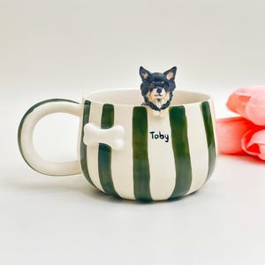 May include: A ceramic mug featuring vertical dark green stripes on a cream background. A three-dimensional black and tan dog figurine is perched on the rim, and a white bone-shaped accent is attached to the side. The name Toby is printed in black.