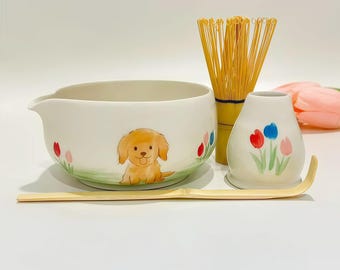 May include: A ceramic matcha bowl with a spout, featuring a golden-brown puppy illustration and small tulips. Includes a bamboo whisk, a ceramic whisk holder with painted tulips, and a long, slender bamboo tea scoop for preparing matcha tea.