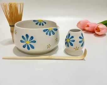May include: A white ceramic matcha tea set with blue and yellow daisy designs. The set includes a bowl, whisk, whisk holder, and scoop. A bamboo whisk and scoop are included. Pink tulips are in the background.