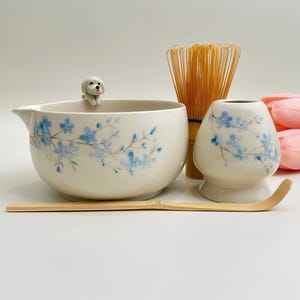 May include: A ceramic matcha bowl with a small white dog figurine on the rim, decorated with blue flowers. Includes a bamboo whisk, a bamboo scoop, and a ceramic whisk holder featuring matching blue floral patterns on a cream-coloured base.