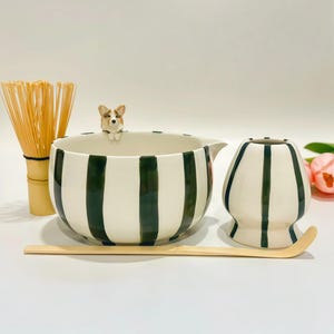 May include: A matcha tea set featuring a ceramic bowl with dark green vertical stripes and a small sculpted corgi figure on the rim, a matching whisk holder, a bamboo whisk, and a long, curved bamboo tea scoop for preparing powdered green tea.