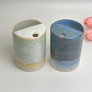 May include: Two ceramic vases with a cylindrical shape. One vase is light green and silver, the other is blue and navy. Each has a diagonal opening and a small hole. The vases are on a white surface.