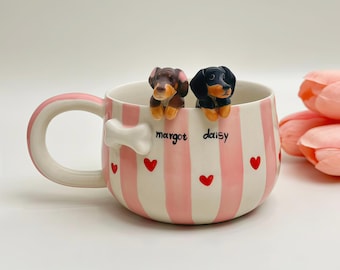 May include: A ceramic mug featuring vertical pink and white stripes, small red hearts, and a white bone-shaped handle accent. Two sculpted dachshund figurines, one brown and one black, peek over the rim. The names Margot and Daisy are printed in black text.