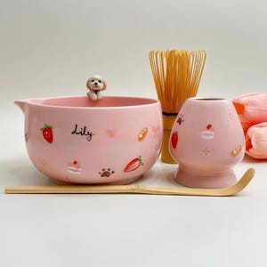 May include: A pink ceramic matcha bowl with a small dog figurine on the rim, featuring the name Lily and illustrations of strawberries, cakes, and paw prints. Includes a matching pink whisk holder, a bamboo whisk, and a bamboo tea scoop.