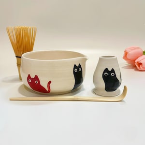 May include: A ceramic matcha tea set featuring a white bowl and a whisk holder, both decorated with black and red cat illustrations. The set includes a light-colored bamboo whisk and a matching bamboo tea scoop, arranged on a plain surface.