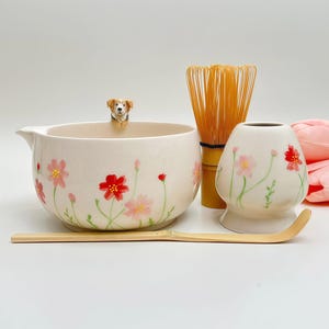 Könnte beinhalten: Eine Matcha-Schale aus Keramik mit einer kleinen Hundefigur am Rand, verziert mit rosa und roten Blumen. Enthält einen Bambusbesen, einen Bambuslöffel und einen passenden Keramikhalter, alle mit handgemalten Blumenmotiven in sanften Farben.