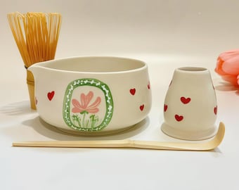 May include: A ceramic matcha bowl with a pink flower illustration in a green frame and small red hearts, a bamboo whisk, a bamboo scoop, and a ceramic whisk holder decorated with red hearts, arranged on a plain white surface.
