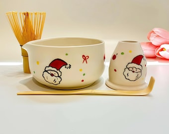 May include: A ceramic matcha bowl and whisk holder featuring a hand-painted Santa Claus motif with red and green confetti accents. The set includes a light-colored bamboo matcha whisk and a long, slender bamboo tea scoop for preparing matcha tea.