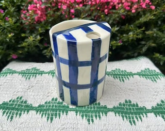 Puede incluir: Un portacepillos de cerámica blanca con un patrón de cuadros azules. El soporte tiene forma rectangular con una parte superior curva y un agujero para un cepillo de dientes. El fondo presenta flores rosas y moradas y una tela estampada blanca y verde.