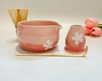 May include: A pink ceramic matcha bowl with a pouring spout and a matching pink ceramic whisk holder, both decorated with white flower motifs. A bamboo whisk and a bamboo tea scoop are included in this set, alongside two soft pink tulip flowers.