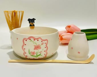 May include: A cream-colored ceramic matcha bowl featuring a hand-painted pink floral frame with tiny red hearts, a small sculpted dog figure on the rim, a bamboo whisk, a bamboo scoop, and a matching cream-colored ceramic whisk holder with pink dots.