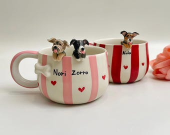 May include: Two ceramic mugs featuring 3D dog figurines on the rims. The front mug has pink stripes, small red hearts, and the names Nori and Zorro. The back mug has red stripes, small red hearts, and the name Nala. A white bone shape is on the handle.