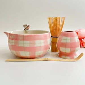 Könnte beinhalten: Ein Matcha-Teeset bestehend aus einer rosa-weiß karierten Keramikschale mit einer kleinen Hundefigur am Rand, einem Bambusbesen, einem Bambuslöffel und einem rosa-weiß karierten Keramikhalter für den Besen auf einer weißen Fläche.