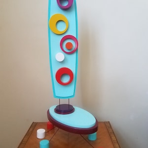 May include: A turquoise blue abstract sculpture with a tall, thin, oval-shaped base and a vertical, rectangular form with colorful circles. The sculpture is made of wood and painted in various colors, including turquoise, purple, yellow, red, orange, and white.