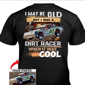 May include: Black t-shirt with a graphic of a dirt racing car, the number 67, and the text "I MAY BE OLD BUT I WAS A DIRT RACER WHEN IT WAS COOL." A skeleton is also featured in the design. A smaller image of the car is in the bottom left.