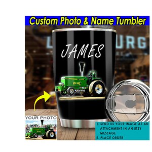 May include: A black and stainless steel tumbler with a custom photo and name. The tumbler features a green tractor pulling a trailer. The text "JAMES" is printed on the tumbler. The tumbler is shown with a lid and a straw.