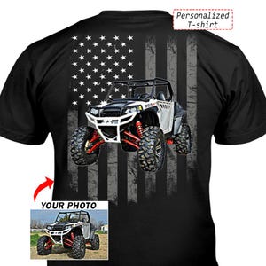 May include: Black t-shirt featuring a graphic of an off-road vehicle with red suspension and large tires, set against a distressed American flag design. The text "Personalized T-shirt" is at the top. A smaller image of the vehicle is at the bottom.