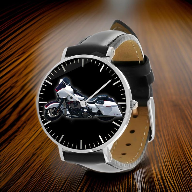 May include: A wristwatch featuring a black leather strap and a round silver-tone case. The watch face displays a black background with a white and black touring motorcycle graphic, white hour markers, and three silver-tone hands for timekeeping.