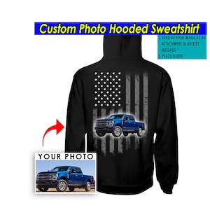 Puede incluir: Sudadera con capucha negra con un diseño de bandera estadounidense desgastado que presenta una camioneta azul. La sudadera se puede personalizar con una foto de tu propia camioneta.