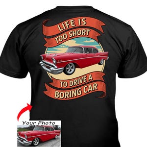 Może przedstawiać: Czarna koszulka z grafiką czerwonego klasycznego samochodu i napisem "LIFE IS TOO SHORT TO DRIVE A BORING CAR." Projekt zawiera czerwony baner i kremowe tło. Mały obrazek samochodu znajduje się na dole.