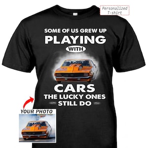 May include: Black t-shirt with the text "SOME OF US GREW UP PLAYING WITH CARS THE LUCKY ONES STILL DO." An orange muscle car graphic is centered on the shirt. A smaller image of the car is in the lower left corner.