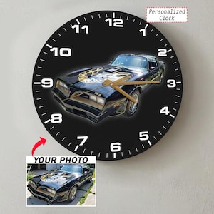 Op de afbeelding: Een ronde wandklok met een zwarte wijzerplaat, witte cijfers en gouden wijzers. Het midden toont een zwarte Pontiac Firebird Trans Am met een gouden motorkapsticker. Een klein kader onderaan toont de originele foto met de tekst YOUR PHOTO.