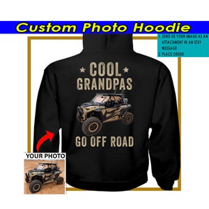 May include: Black hoodie with a graphic of a side-by-side off-road vehicle and the text "COOL GRANDPAS GO OFF ROAD".  The hoodie has a pocket on the front and a hood.  The graphic is in a camouflage pattern.  The text is in a brown font.  There is a red arrow pointing to the area where a photo can be added.  A smaller image of a side-by-side off-road vehicle is shown in the lower left corner of the image.