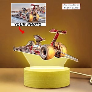 May include: A personalized night light featuring a clear acrylic cutout of a red drag racing car mounted on a cylindrical light-up base. The design includes a printed image of a dragster on a track with the text YOUR PHOTO and Personalized Night Light.