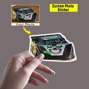 May include: A custom photo sticker featuring a black race car with a skull design on the front. The sticker has a white border and the text "Custom Photo Sticker" in a yellow rectangle.
