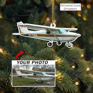 May include: A white and brown aeroplane ornament with the text N7706X on the side, hanging from a gold chain. Below it, a rectangular photo shows the same aeroplane on a grassy field with the text YOUR PHOTO and Personalized Ornament visible.