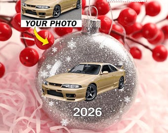 Może przedstawiać: Okrągła, srebrna, brokatowa bombka choinkowa z nadrukowanym zdjęciem złotego samochodu sportowego Nissan Skyline GT-R oraz rokiem 2026 w białym kolorze. Ozdoba posiada srebrną zawieszkę i jest udekorowana grafikami białych płatków śniegu.