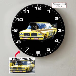 May include: A round wall clock with a black face and white numbers. The center features a yellow drag racing car with a large engine intake. Gold clock hands are positioned over the car. A small inset shows the original photo of the yellow car.