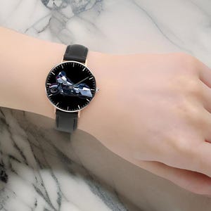 May include: A wristwatch featuring a round face with a black dial and a detailed graphic of a motorcycle. The watch has a thin rose gold-tone metal case, slender hour and minute hands, and a smooth black leather strap with a buckle closure.