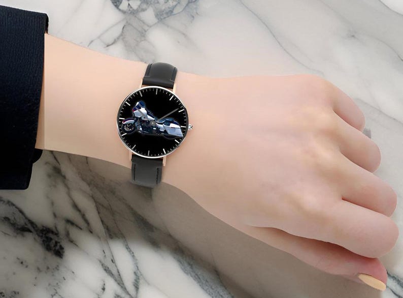 May include: A wristwatch featuring a round face with a black dial and a detailed graphic of a motorcycle. The watch has a thin rose gold-tone metal case, slender hour and minute hands, and a smooth black leather strap with a buckle closure.