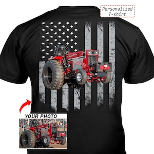 May include: Black t-shirt featuring a red tractor design set against a distressed American flag. The tractor has large tires and the text "Christine" is visible. The shirt also includes the text "Personalized T-shirt" and "YOUR PHOTO".