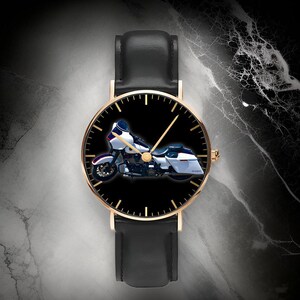 May include: A wristwatch with a black leather strap and a gold-tone circular case. The watch face is black and features a graphic of a white and blue touring motorcycle in the center, with gold-tone hour markers and thin gold-tone hands.