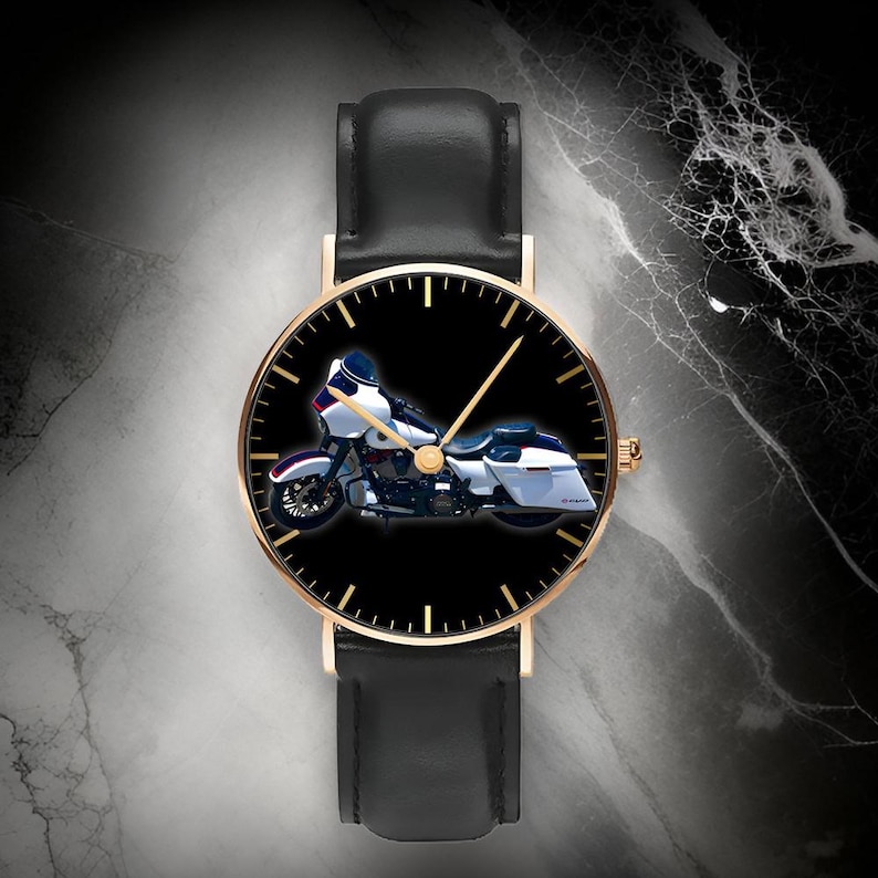 May include: A wristwatch with a black leather strap and a gold-tone circular case. The watch face is black and features a graphic of a white and blue touring motorcycle in the center, with gold-tone hour markers and thin gold-tone hands.