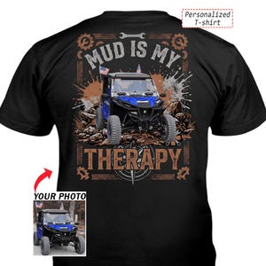 Op de afbeelding: Een zwart t-shirt met een afbeelding van een blauw terreinvoertuig op rotsachtig terrein met modderspatten. De tekst luidt MUD IS MY THERAPY. Een kleiner inzetstuk toont het voertuig met de tekst YOUR PHOTO en Personalized T-shirt.