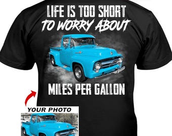 May include: A black t-shirt featuring a graphic of a bright blue vintage pickup truck. The text reads "LIFE IS TOO SHORT TO WORRY ABOUT MILES PER GALLON" in white distressed lettering. A smaller inset box shows the same truck with "YOUR PHOTO" text.